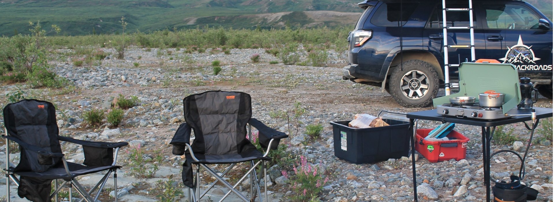 Denali Backroads Overlander vehicle with rooftop tent and camp setup in Alaska wilderness featuring chairs, camp kitchen and essential gear.