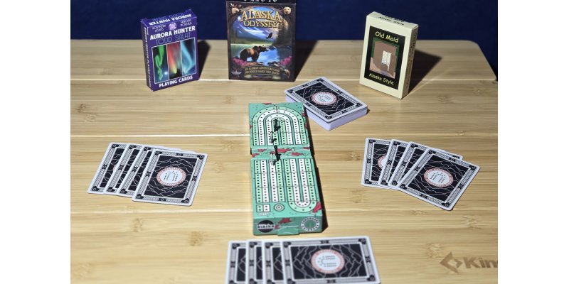 Setup of Alaska-themed games on a wooden table for Denali Backroads Gamers Upgrade: Aurora Hunter Todd Salat Northern Lights playing cards, Alaska Odyssey card game box, Old Maid Alaska Style, custom cribbage board, and spread cards ready for play during cozy Auroralander or Overlander mode adventures