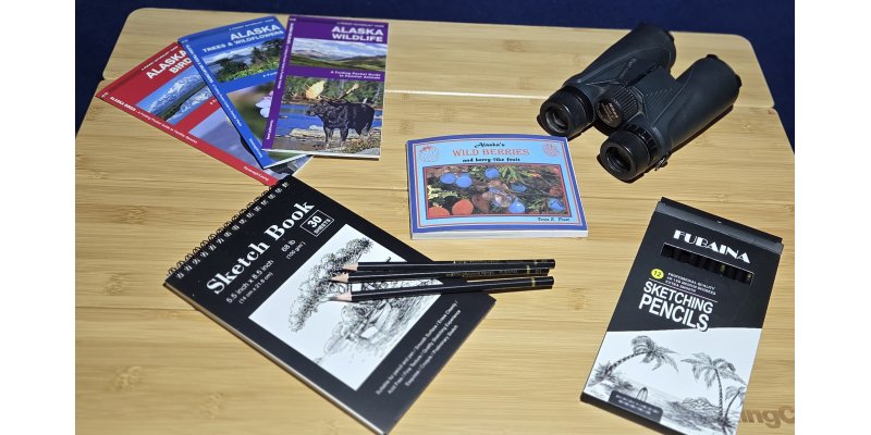 Alaska Naturalist Kit laid out on wooden table including folding pocket field guides to Alaskan birds, trees & wildflowers and wildlife; black spiral sketchbook; Fubaina professional sketching pencils; and compact black binoculars for wildlife observation and nature journaling on Denali Backroads adventures
