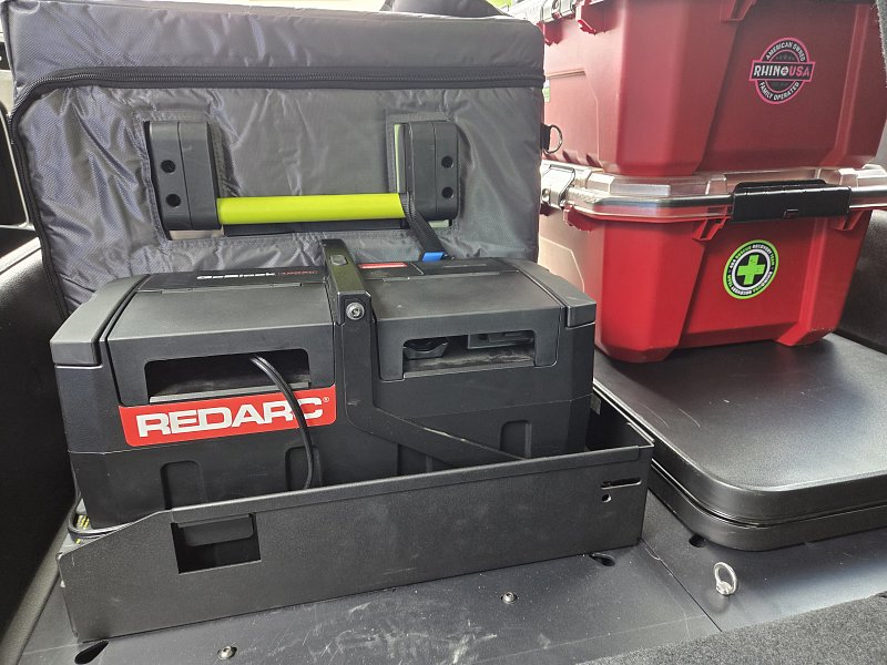 Organized rear cargo area of a Denali Backroads Overlander vehicle showing camp kitchen bins, refrigerator/freezer, cooking gear and fully equipped camping supplies ready for an Alaska adventure.