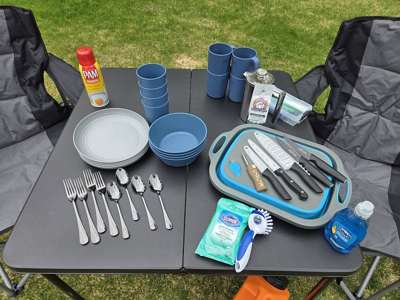 Overlanding camp dining kit with plates, bowls, cups, knives, utensils and outdoor kitchen supplies on a folding table