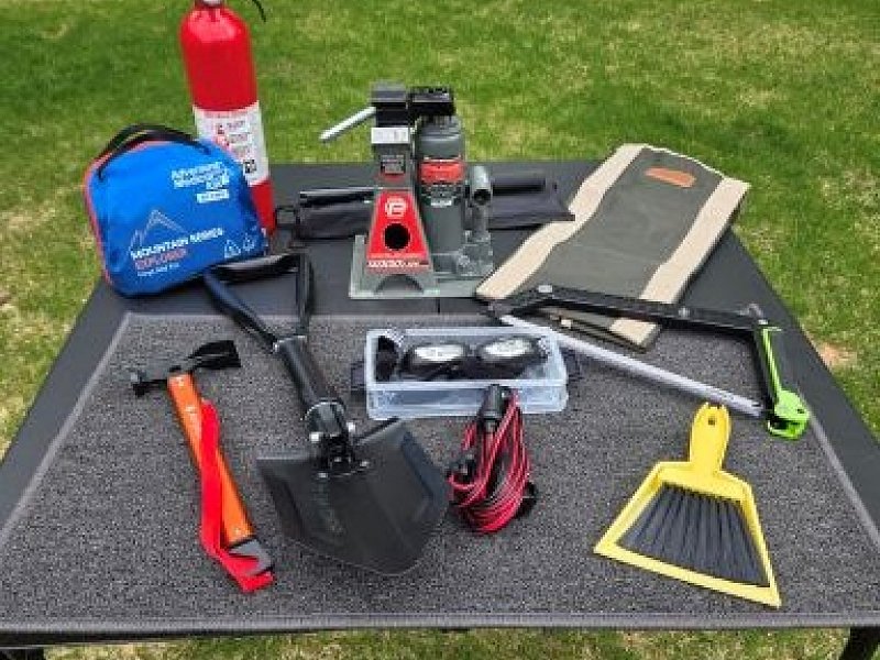 Denali Backroads Overlander safety gear displayed on a table including jack, shovel, saw, fire extinguisher, first aid kit, recovery tools and emergency equipment for off-road travel in Alaska.