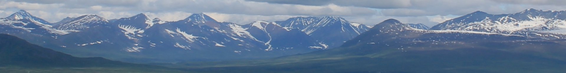 Wide panoramic view of Alaska mountain range with snow-covered peaks and cloudy northern sky.